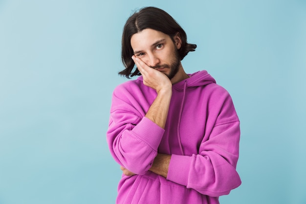 Portrait of a depressed young bearded brunette man wearing hoodie standing isolated over blue wall