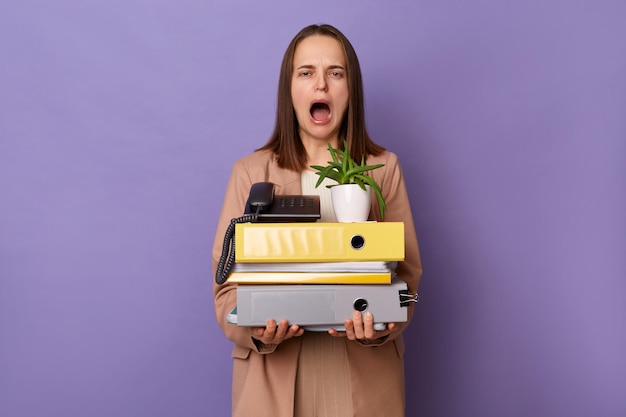 Portrait of despair young adult woman wearing beige jacket\
holding lot of documents folders screaming expressing negative\
emotions posing isolated over lilac background