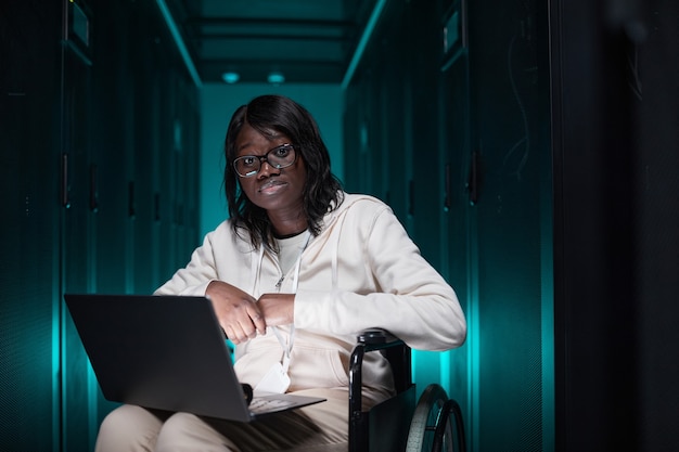 Портрет афро-американской женщины-инвалида, использующей ноутбук и смотрящей в камеру во время работы в серверной, доступная возможность трудоустройства, место для копирования