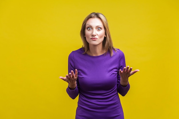 Photo portrait of disappointed unhappy woman in elegant tight purple dress standing with raised hands asking what do you want, angry face. indoor studio shot isolated on yellow background