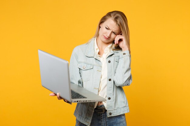 Portrait of displeased crying young woman in denim casual clothes holding, using laptop pc computer isolated on yellow orange wall background in studio. People lifestyle concept. Mock up copy space.