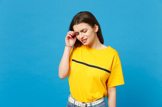 Portrait of displeased crying young woman in vivid casual clothes looking down, keeping hand near face isolated on bright blue background wall in studio. People lifestyle concept. Mock up copy space.