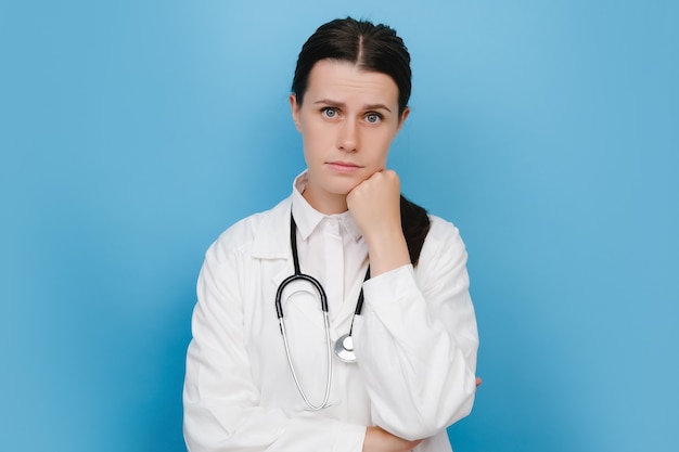 Portrait of displeased offended young doctor woman keeps hands half crossed, holds chin, has angry expression, being deep in thoughts, dressed in uniform and stethoscope, models over blue background