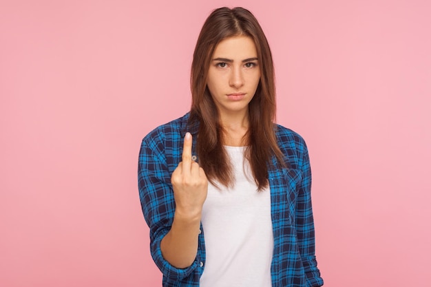 Portrait of displeased vulgar girl in checkered shirt showing\
middle finger impolite rude gesture of disrespect and hate looking\
angrily at camera indoor studio shot isolated on pink\
background