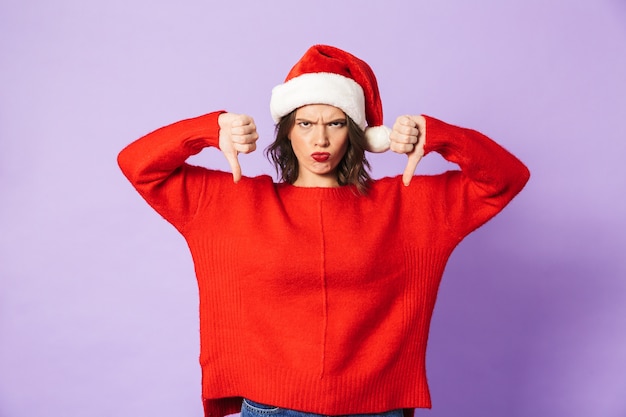 Portrait of a displeased young woman wearing christmas hat isolated over purple wall showing thumbs down gesture.