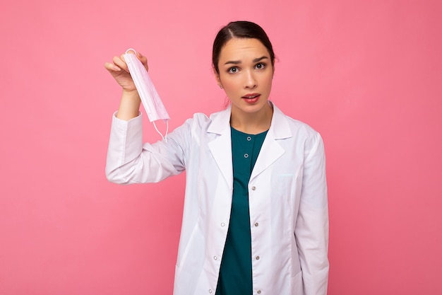 ピンクの背景に分離された医療マスク立っている白いコートで不満の魅力的な若い女性医師の肖像画。