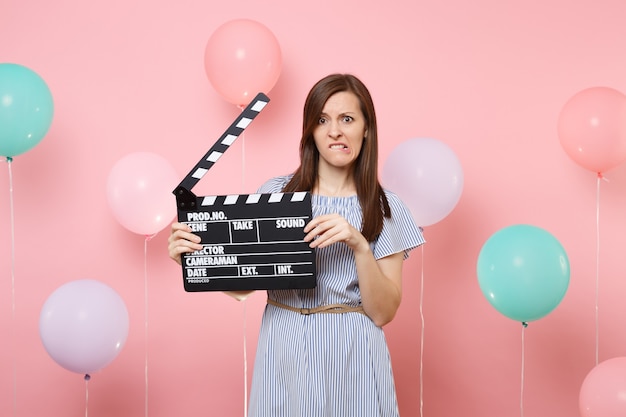 Portrait of dissatisfied concerned woman in blue dress biting lips holding classic black film making clapperboard on pastel pink background with colorful air baloons. Birthday holiday party concept.