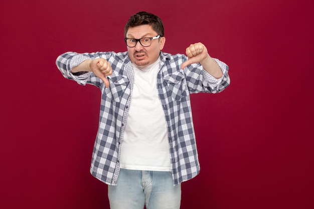 Portrait of dissatisfied handsome middle aged businessman in casual checkered shirt and eyeglasses standing and looking at camera with dislike thumbs down. studio shot, isolated on dark red background