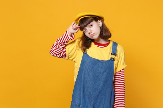 Portrait of dissatisfied sad girl teenager in french beret, denim sundress looking aside isolated on yellow wall background in studio. People sincere emotions, lifestyle concept. Mock up copy space.