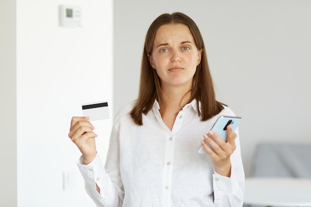 明るい部屋で屋内に立って、携帯電話とクレジットカードを手に持って、カメラを見て、オンラインショッピング、キャッシュバックの白いシャツを着ている不満のある女性の肖像画。