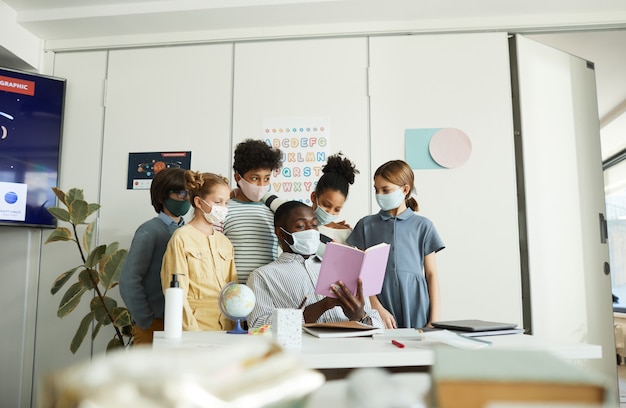 Ritratto di un gruppo eterogeneo di bambini con insegnante maschio che indossa maschere in classe scolastica, misure di sicurezza covid, spazio copia