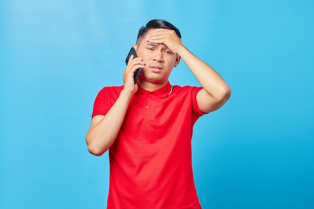 Portrait of dizzy asian man talking on smartphone and holding head with palm isolated on blue background