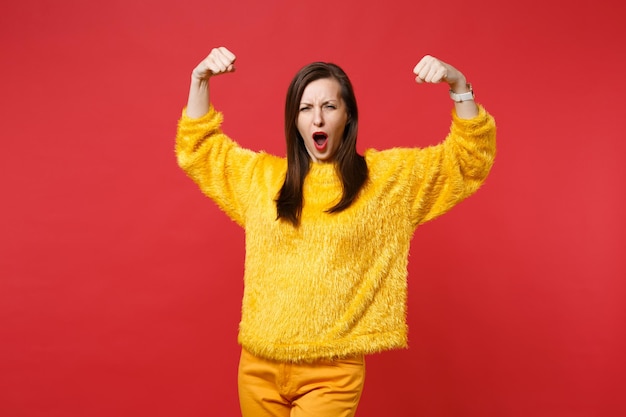 Portrait of dizzy funny crazy young woman in yellow fur sweater showing biceps muscles isolated on bright red wall background in studio. People sincere emotions, lifestyle concept. Mock up copy space.