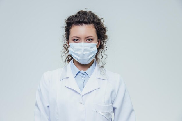 Portrait of a doctor of a beautiful and young woman In a white robe there is a gray background Dressed in a white face mask
