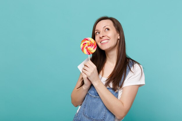 Portrait of dreamful smiling young woman in denim clothes looking up holding colorful round lollipop isolated on blue turquoise wall background in studio. People lifestyle concept. Mock up copy space.
