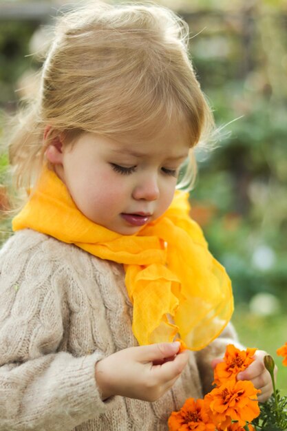 Photo portrait of dreaming little blonde girl in yellow scarf looking at orange flowers in her hand