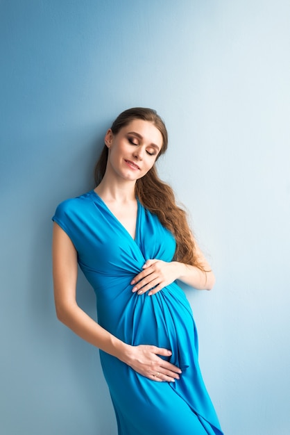 Portrait of a dreaming pregnant woman in a blue dress on a blue background