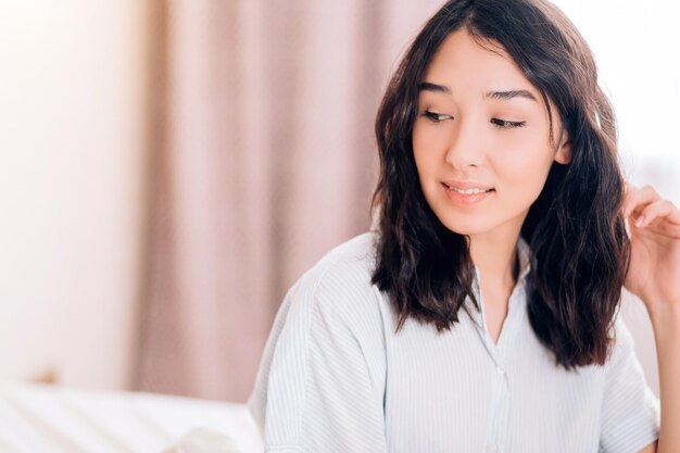 Portrait of dreamy beautiful caucasian girl with natural makeup, in pajamas sitting in bed in hotel, smiling looking away, remembering date with her boyfriend. Morning time, relaxation. Copy space.