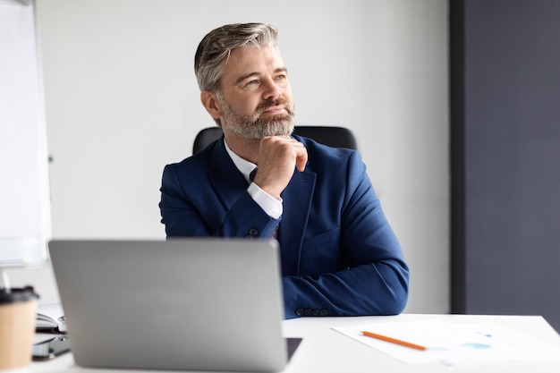 Portrait Of Dreamy Mature Businessman Sitting At Desk With Laptop In Office