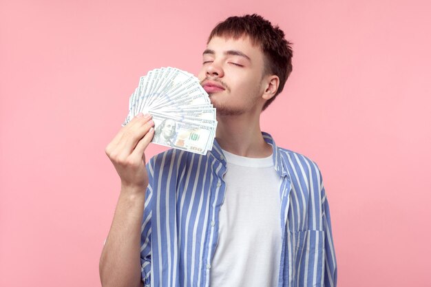 Portrait of dreamy rich brownhaired man with small beard and mustache in casual striped shirt smelling money dreaming with closed eyes about profit indoor studio shot isolated on pink background