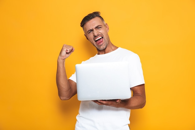 Portrait of ecstatic guy 30s in white t-shirt holding and using silver laptop isolated on yellow