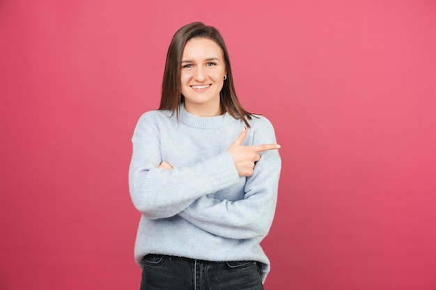 Portrait of an ecstatic woman is pointing aside while smiling at the camera Studio shot over pink background