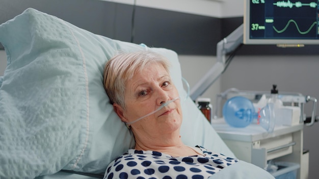 Portrait of elder patient with oxygen tube laying in hospital ward bed. Woman with illness looking at camera while waiting for healthcare assistance in emergency room for intensive care