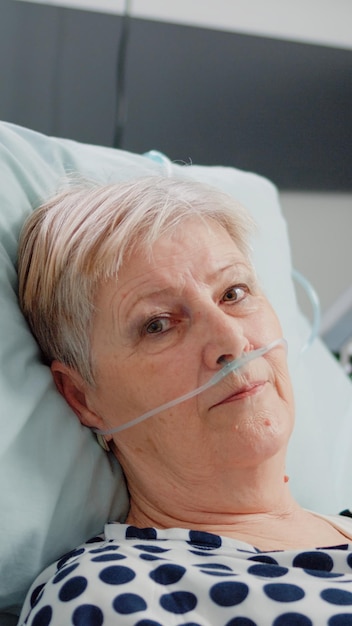 Portrait of elder patient with oxygen tube laying in hospital ward bed. Woman with illness looking at camera while waiting for healthcare assistance in emergency room for intensive care