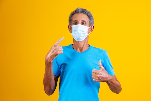 Portrait of elderly afro with surgical medical mask standing in blue shirt with thumb up making thumbs up ok while pointing to mask on his face