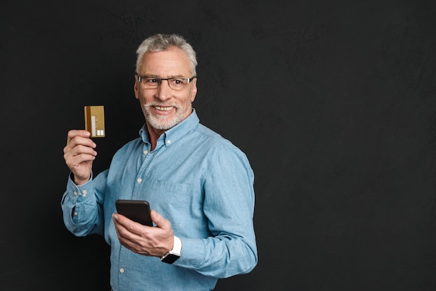 Portrait of elderly businessman 70s with grey hair and beard holding credit card and smartphone for paying online, isolated over black wall