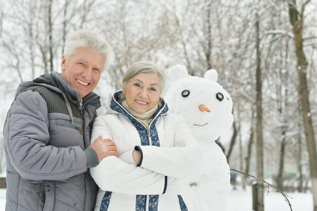 Portrait of elderly couple making snowman and shows thumbs up in winter