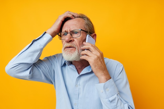 Portrait elderly man in a blue shirt and glasses talking on the phone yellow background