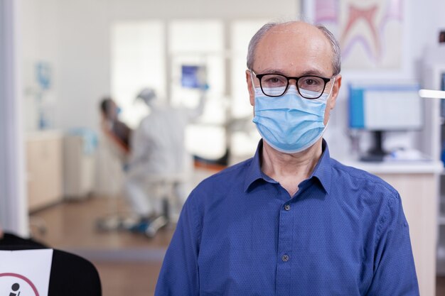Портрет пожилого мужчины в стоматологическом кабинете, глядя в камеру в маске для лица, сидя на стуле в зале ожидания стоматологической клиники