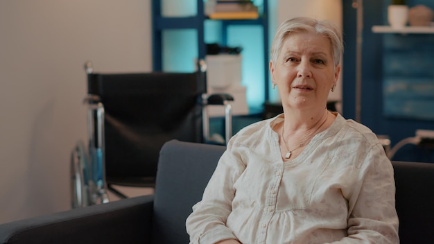 Portrait of elderly woman looking at camera in living room. Senior person with chronic disability enjoying retirement and smiling. Old adult sitting next to wheelchair for transportation.