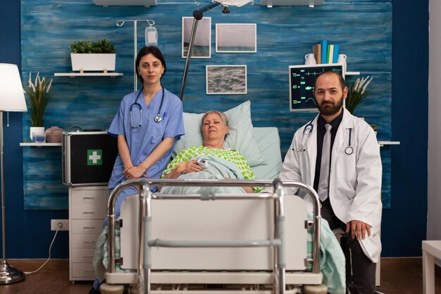 Portrait of elderly woman lying in nursing home bed with\
medical team attending her in the room. doctor and nurse\
specializing in elderly care, health assistance service.