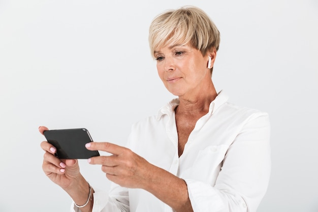 portrait of elegant adult woman with short blond hair wearing earpods and holding cellphone isolated over white wall in studio