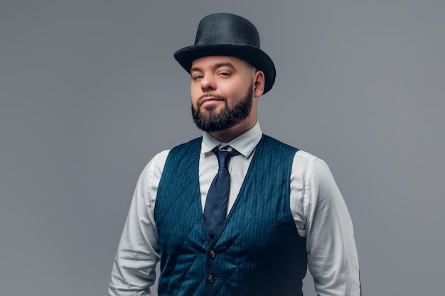 Photo portrait of elegant bearded male dressed in a waistcoat and a cylinder hat isolated on grey background.