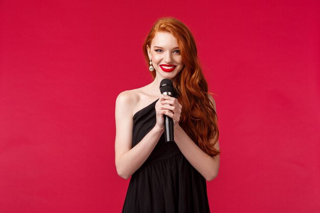 Portrait of elegant beautiful young woman with red long hair wearing evening dress, holding microphone, performer singing for clients in restaurant, smiling sensually, red wall