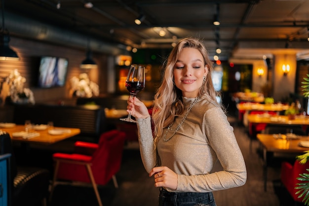 Portrait of elegant blonde woman holding glasses of red wine standing in restaurant with luxury interior with dark interior looking away