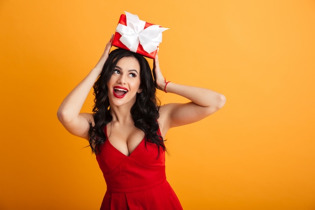 Portrait of elegant lovely woman 20s in red dress taking pleasure in receiving present box on birthday, isolated over yellow wall