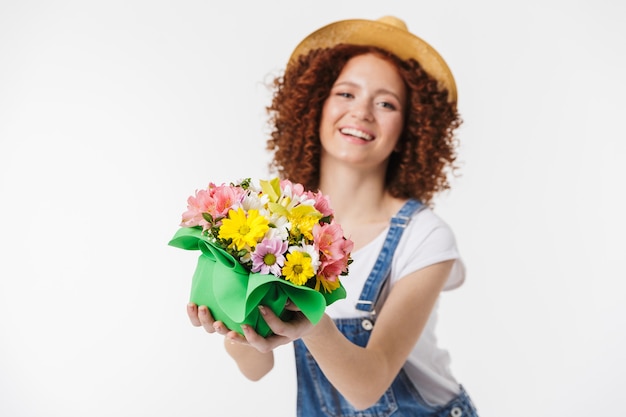 Portrait of elegant redhead curly woman 20s wearing summer straw hat smiling and holding flower box isolated over white wall