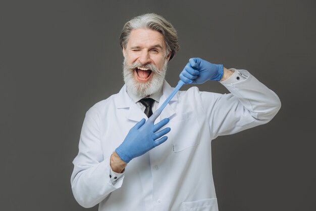 Portrait of emotional doctor on gray wall. Gray-haired senor doctor dressed in white coat and blue gloves isolated on gray background.