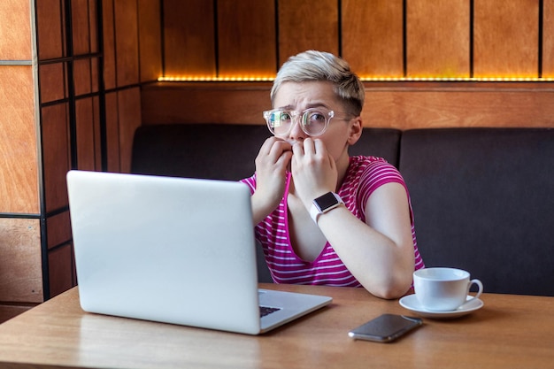 Portrait of emotional nervous young girl with blonde short hear in pink t-shirt and eyeglasses is sitting in cafe, working alone and biting nail herself. Indoor, looking at camera, lifestyle