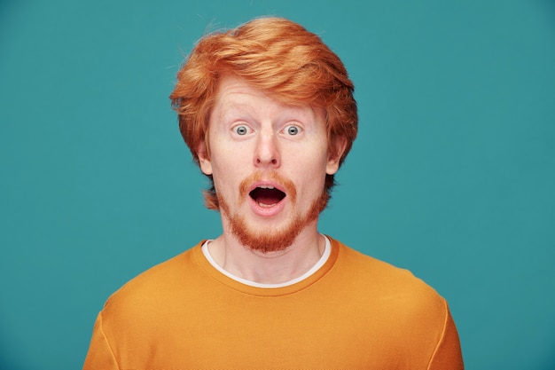 Portrait of emotional young red-bearded man surprised by news keeping mouth open on blue