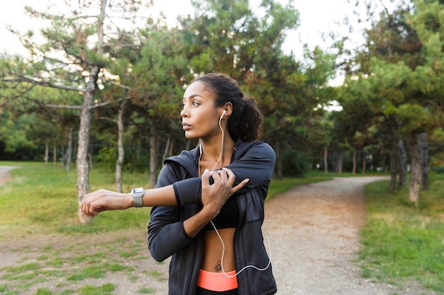 Portrait of energetic african american woman 20s wearing black tracksuit doing exercises, and stretching her body in green park