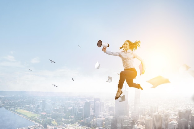 Portrait of energetic businesswoman with loud speaker jumping in open air. Mixed media