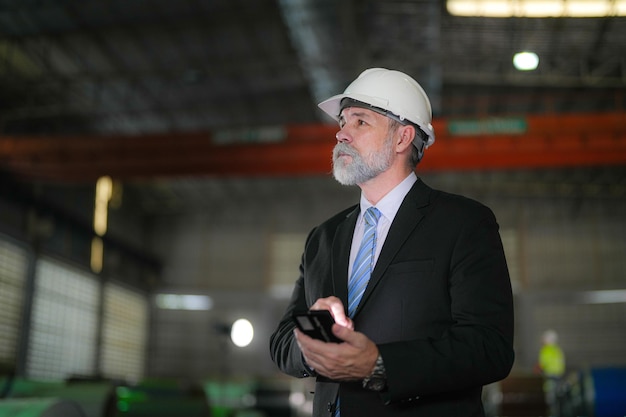 Portrait engineer Senior boss standing confident and holding smartphone in industry factory Concept of smart industry operation and maintenance older engineer with beard mustache on face standing