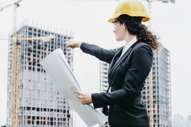 A portrait of engineer woman looking at construction. Woman architect with blueprint in hand and yellow helmet with select focus. Concept Engineer and business work.