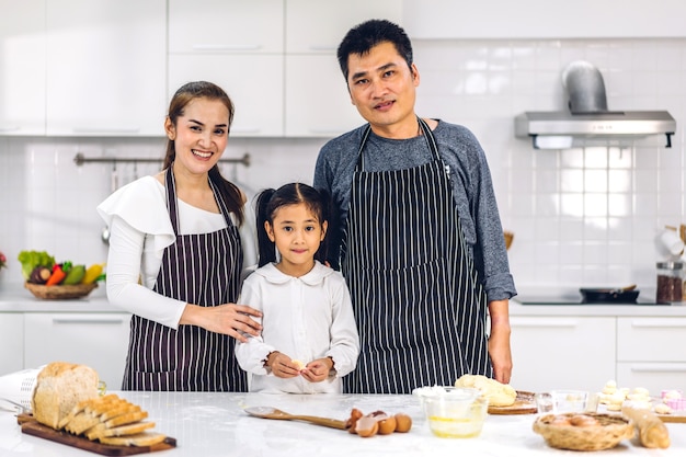 Ritratto di godere di amore felice asiatico padre di famiglia e madre con la piccola ragazza asiatica figlia bambino divertendosi cucinando insieme a cuocere i biscotti e gli ingredienti della torta sul tavolo in cucina