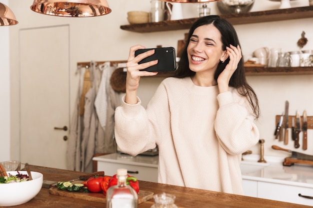 Portrait of european brunette woman taking selfie photo on smartphone while cooking green salad with vegetables in stylish kitchen at home